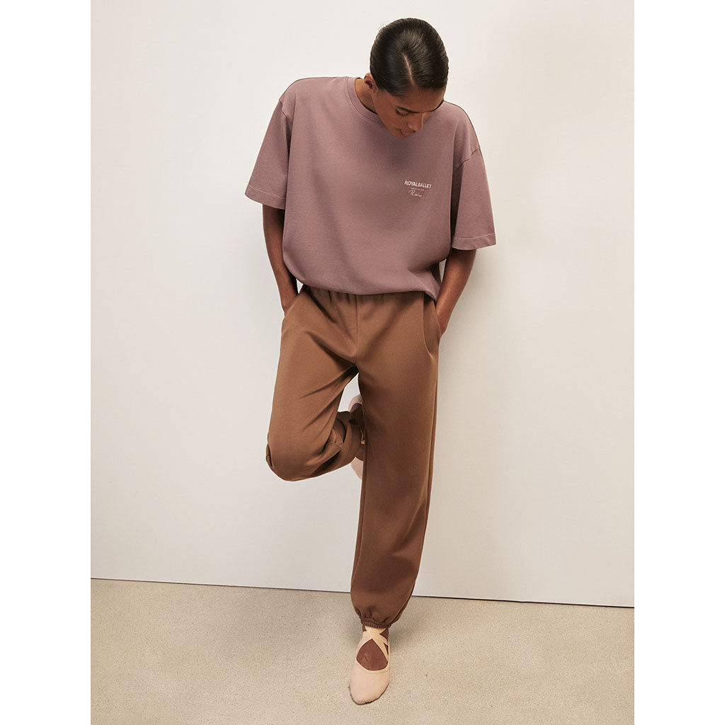 Person wearing a rose t-shirt and brown pants  and ballet pumps against a white wall.