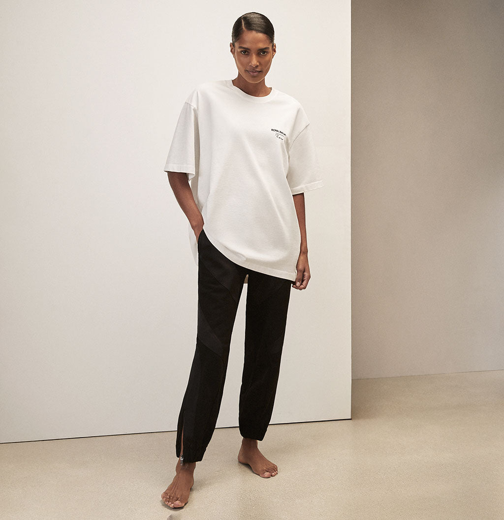 Person wearing a white oversized t-shirt with Royal Ballet Reiss on left chest and black pants against a minimalistic background