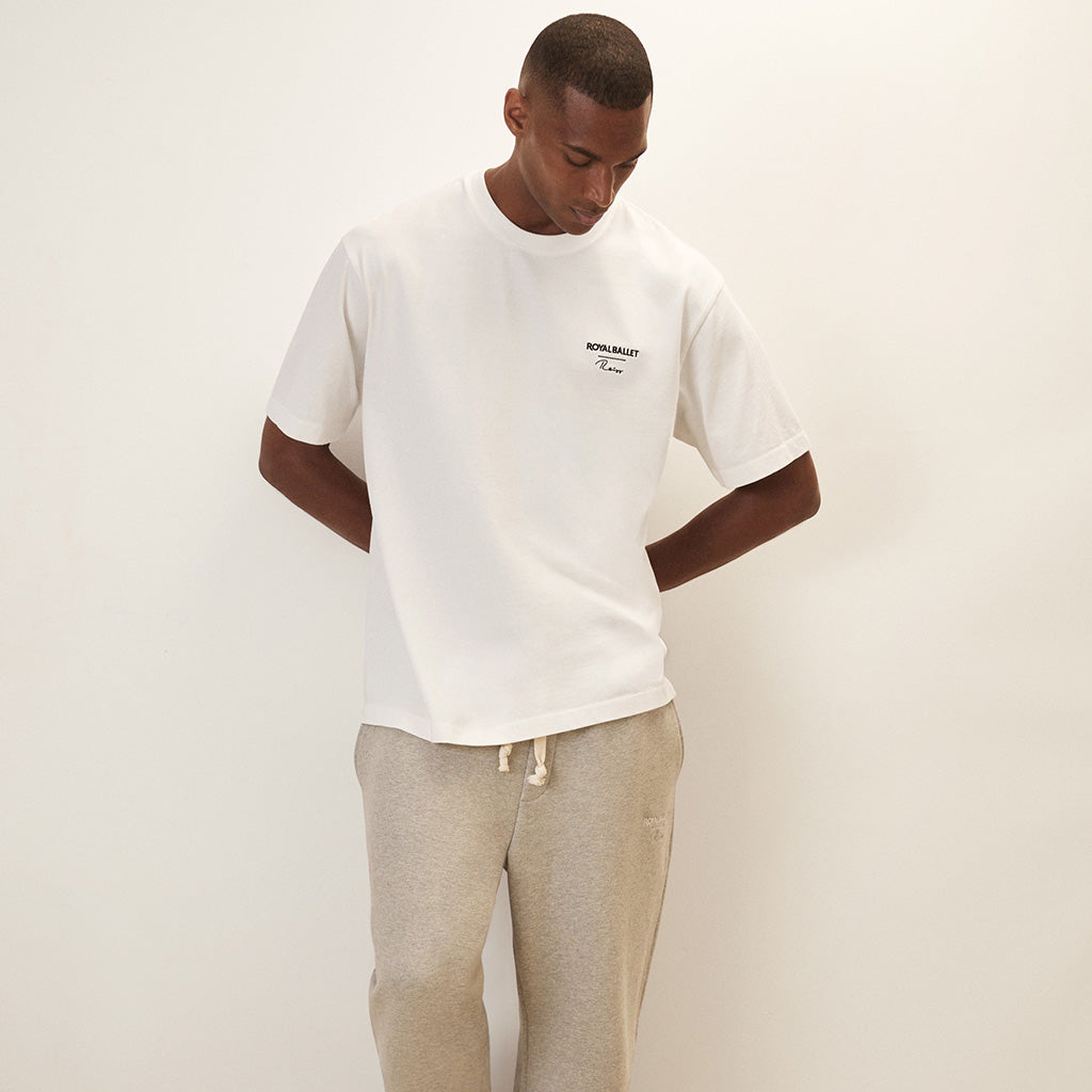 Man wearing a white  t-shirt with Royal Ballet Reiss on left chest and grey pants leaning against a white wall