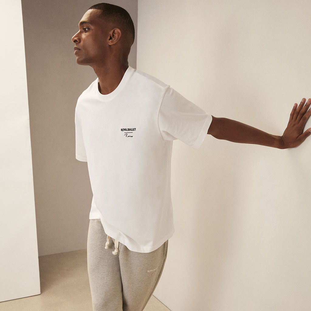 Man wearing a white  t-shirt with Royal Ballet Reiss on left chest and grey pants against a minimalistic background