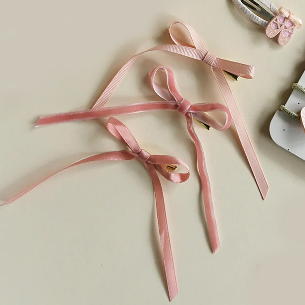 A set of 3 Long Ribbon Bow Clips on a white card with black dots, against a white background. The three bow clips are all pink, with the top being the lightest, the middle being slightly darker, and the bottom being made from velvet. They are all tied into a bow with long tails hanging below the loops. 