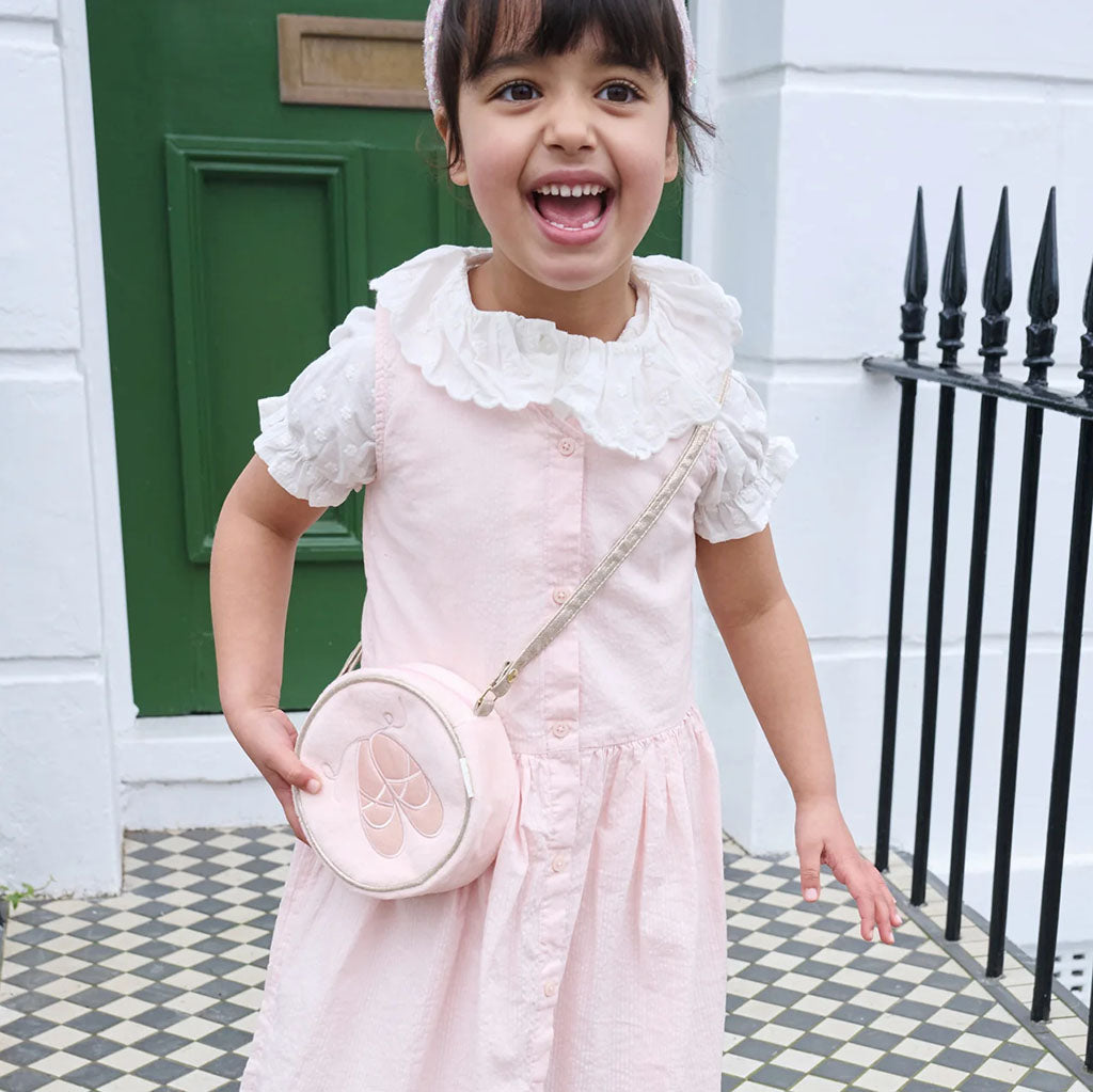 A round, childrens light pink fabric crossbody bag featuring an embroidered ballet shoe design in the centre, complete with a matching adjustable shoulder strap and a small gold-toned zipper pull.