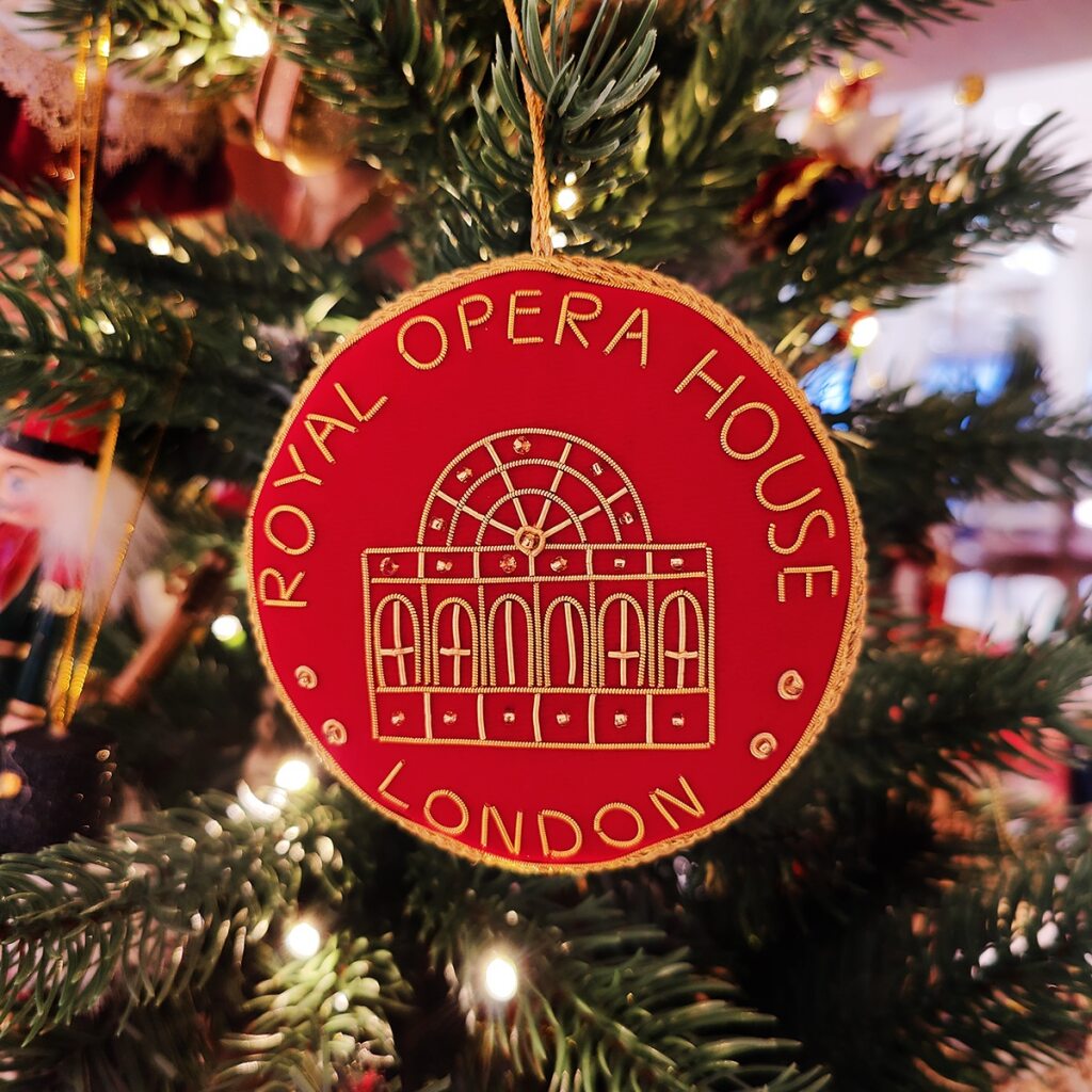 Red velvet roundel decoration with gold text 'Royal Opera House London ' and the facade of the building on a white background