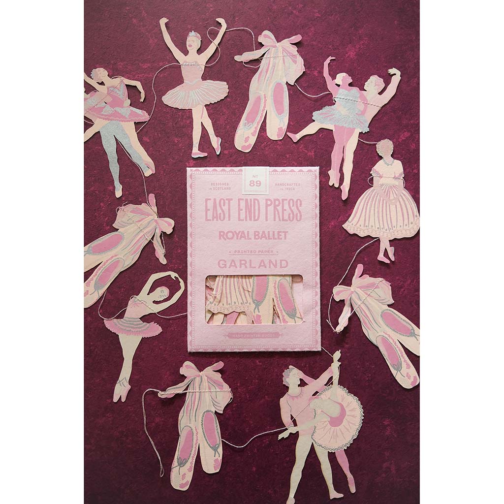 A package of East End Press Royal Ballet ballerina paper garland, surrounded by several cutout ballerina figures in pink and cream tulle dresses on a textured dark magenta background.