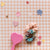 A Set of four hair scrunchies on a piece of white card with black dots, set against a white background. The top scrunchie is blue with pink, white, and yellow flowers, the second scrunchie is white with pink flowers, the third scrunchie is a coral coloured gingham pattern, and the final scrunchie is white with yellow flowers. 