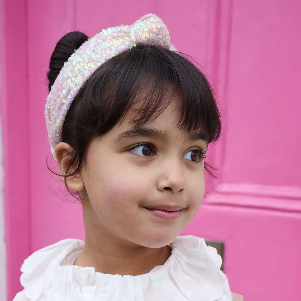 A young girl with dark hair in a high bun wearing a sparkling iridescent sequin pink  headband, set against a vibrant pink door background.