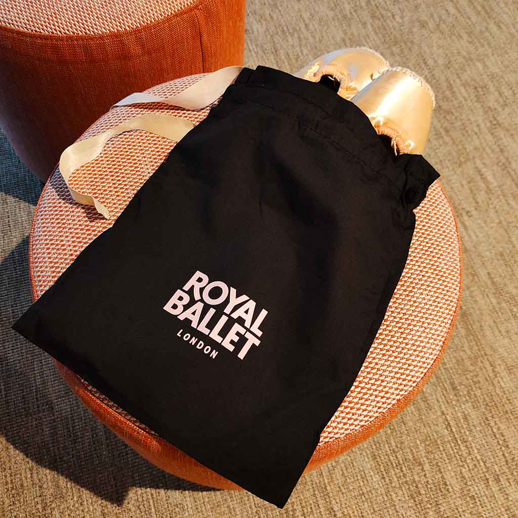 Pointe shoes in a black drawstring bag with 'Royal Ballet London' text on a fabric stool.
