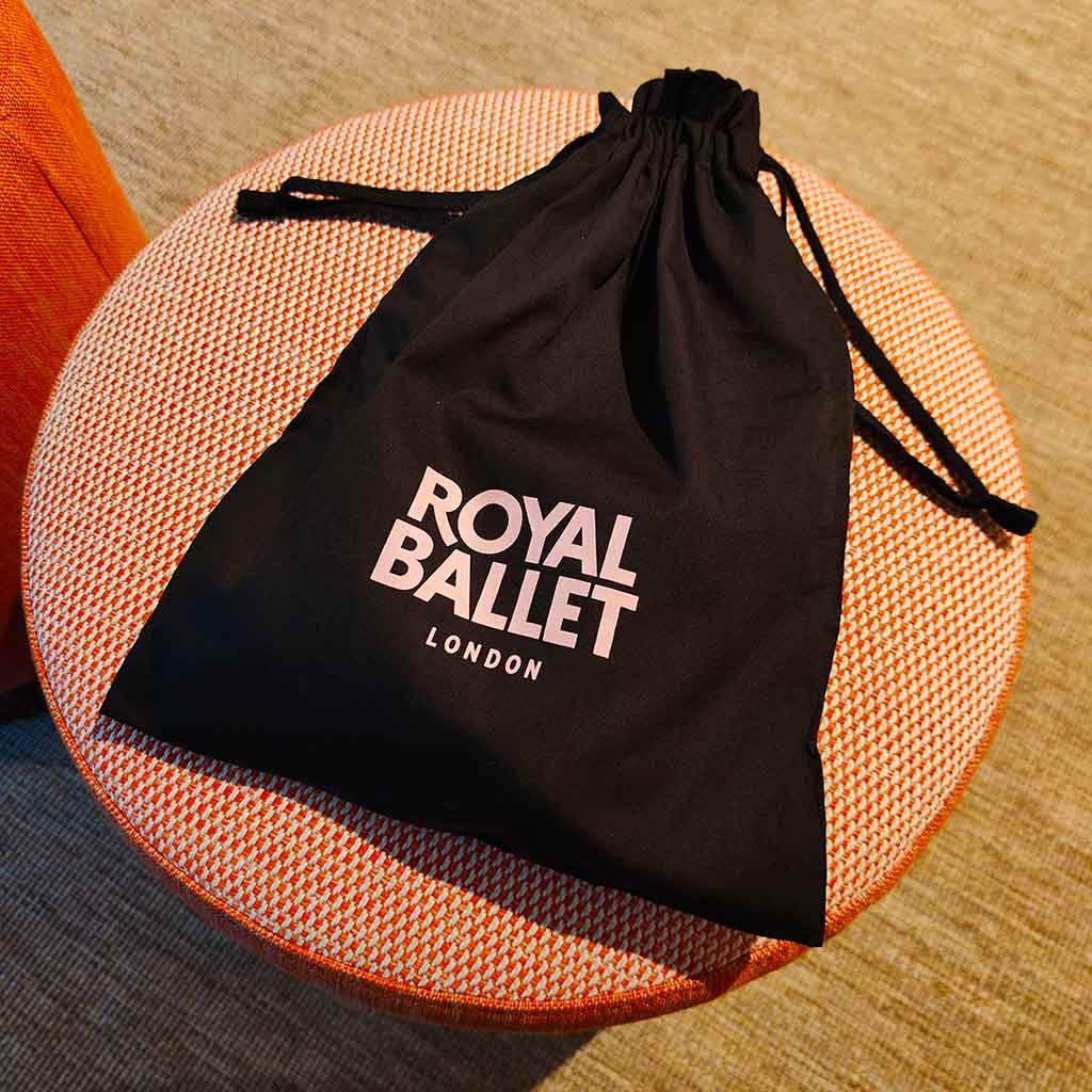 Pointe shoes in a black drawstring bag with 'Royal Ballet London' text on a fabric stool.