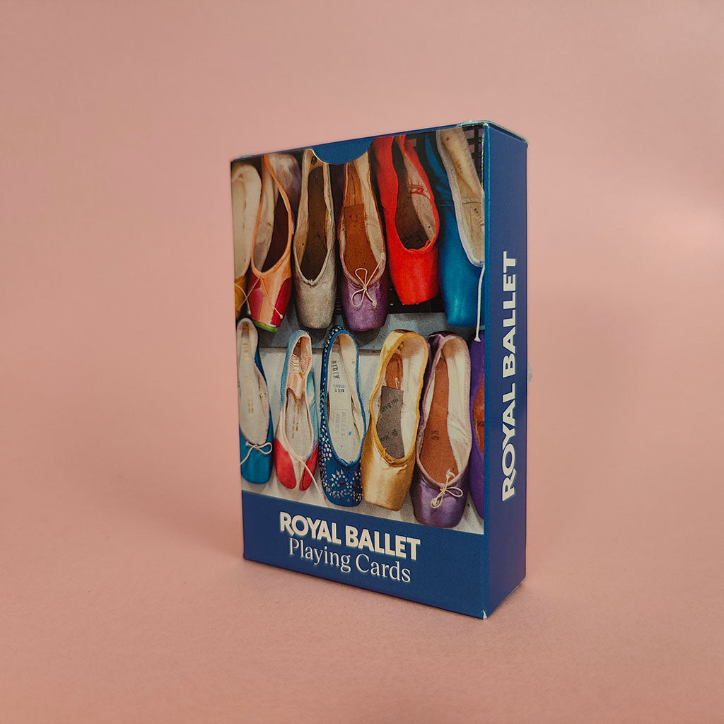 A blue box of Royal Ballet Playing Cards standing upright on a dusty pink background. The front of the box features a colourful photograph of ballet pointe shoes, and the side displays the brand name 'ROYAL BALLET' in white text.