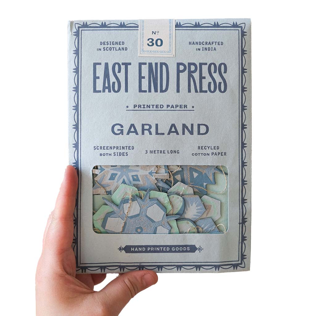A hand holding a package of East End Press Garland Screenprinted Paper. The packaging is a light blue-gray colour with navy blue text and border details, and features a clear window showing the patterned paper inside.
