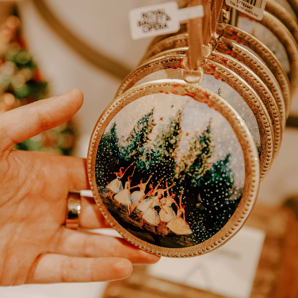 We can see a Snowflakes tin Decoration with what looks like many more hanging directly behind it. A hand is holding the front decoration and tilting it to face the viewer. The background is out of focus but the composition suggests that these decorations are on display in a shop. 