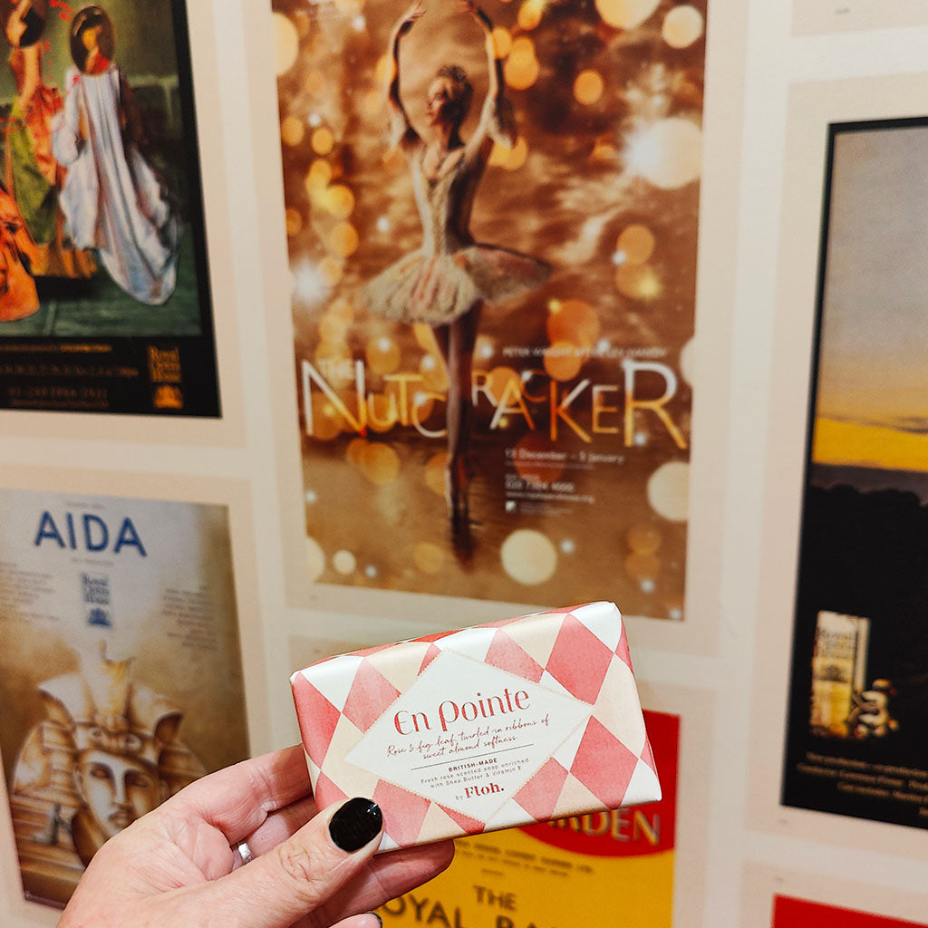 A hand with black nail polish holds a pink and white checkered bar of En Pointe soap in front of a wall of framed posters, including a Nutcracker poster with Darcey Bussell en pointe.