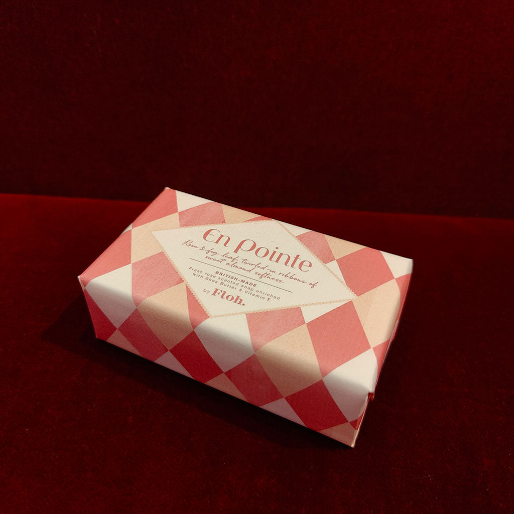 A wrapped bar of soap with pink and cram harlequin diamond pattern, featuring a central cream label that reads 'En pointe' and 'FLOH', photographed on a dark red velvet-like surface.