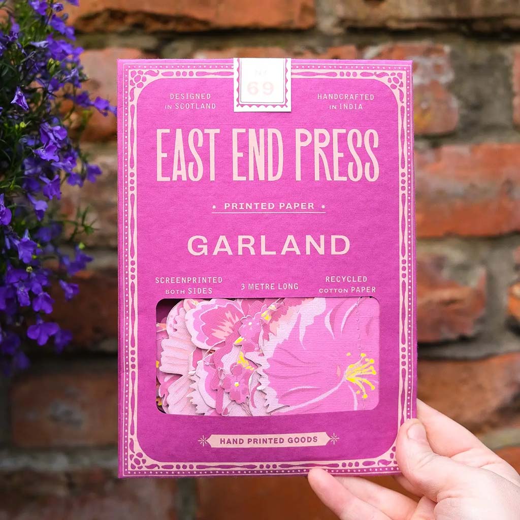 Purple packaging of East End Press Garland with floral design, held by a hand against a rustic background.