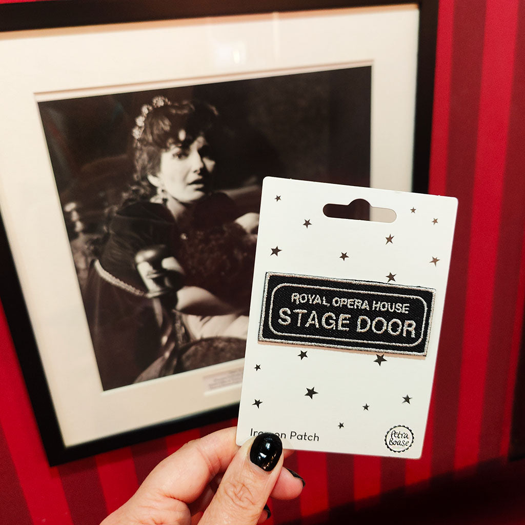 Hand holding a 'Royal Opera House Stage Door' patch in front of a framed black and white photo of Maria Callas on a red striped wall.