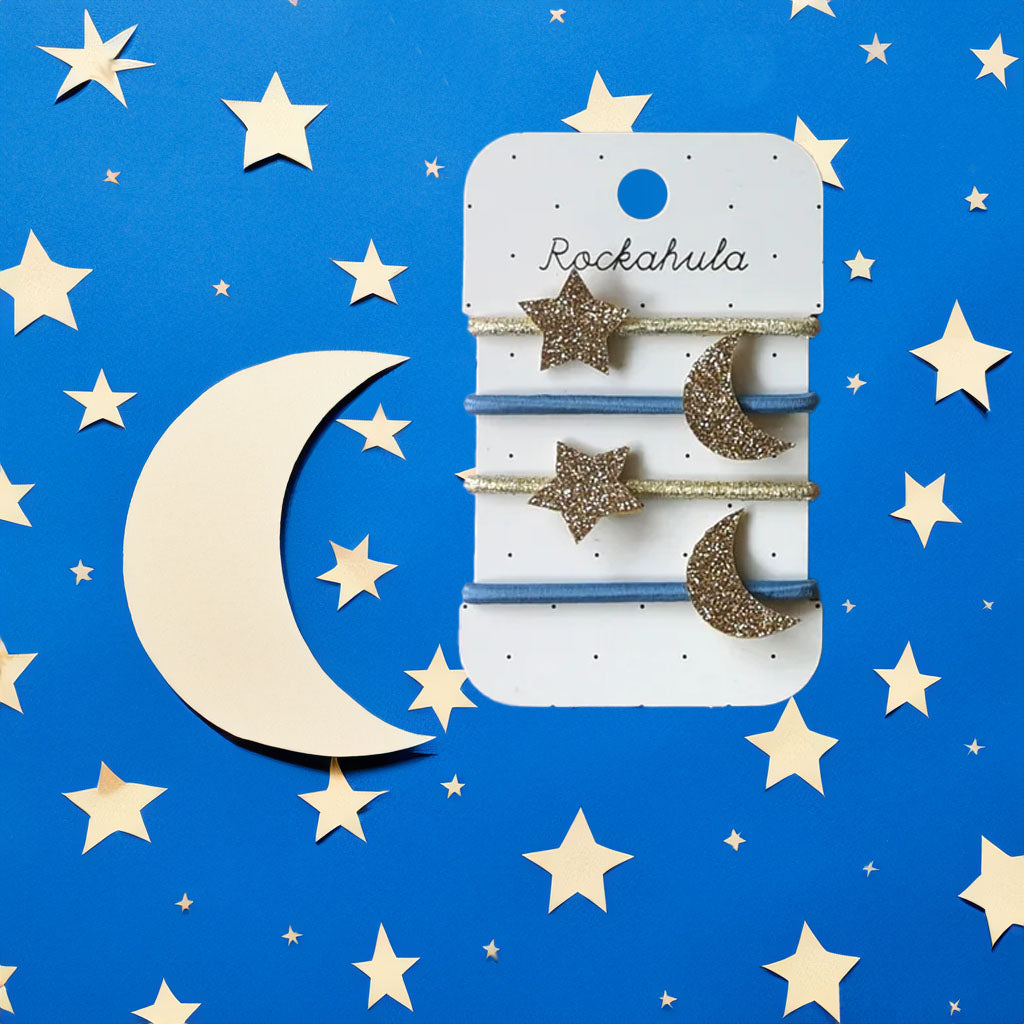 A set of Starry Skies Hair Ties on a white card with black dots against a white background. There are four elastic ties, two gold and two blue. The gold ties have a glittery star attached and the blue ties have a glittery crescent moon attached. 