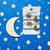 A set of Starry Skies Hair Ties on a white card with black dots against a white background. There are four elastic ties, two gold and two blue. The gold ties have a glittery star attached and the blue ties have a glittery crescent moon attached. 
