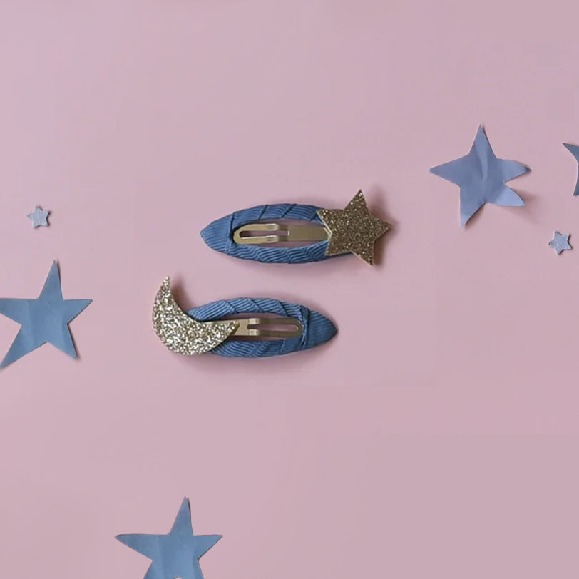 A pair of Starry Skies hair clips on a white card with black dots against a white background. There is a pink fabric hook attached to the top of the card. The hair clips have a blue fabric body, the top one has a glittery moon attached to the left side, and the bottom has a glittery star attached to the right side. 
