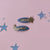 A pair of Starry Skies hair clips on a white card with black dots against a white background. There is a pink fabric hook attached to the top of the card. The hair clips have a blue fabric body, the top one has a glittery moon attached to the left side, and the bottom has a glittery star attached to the right side. 