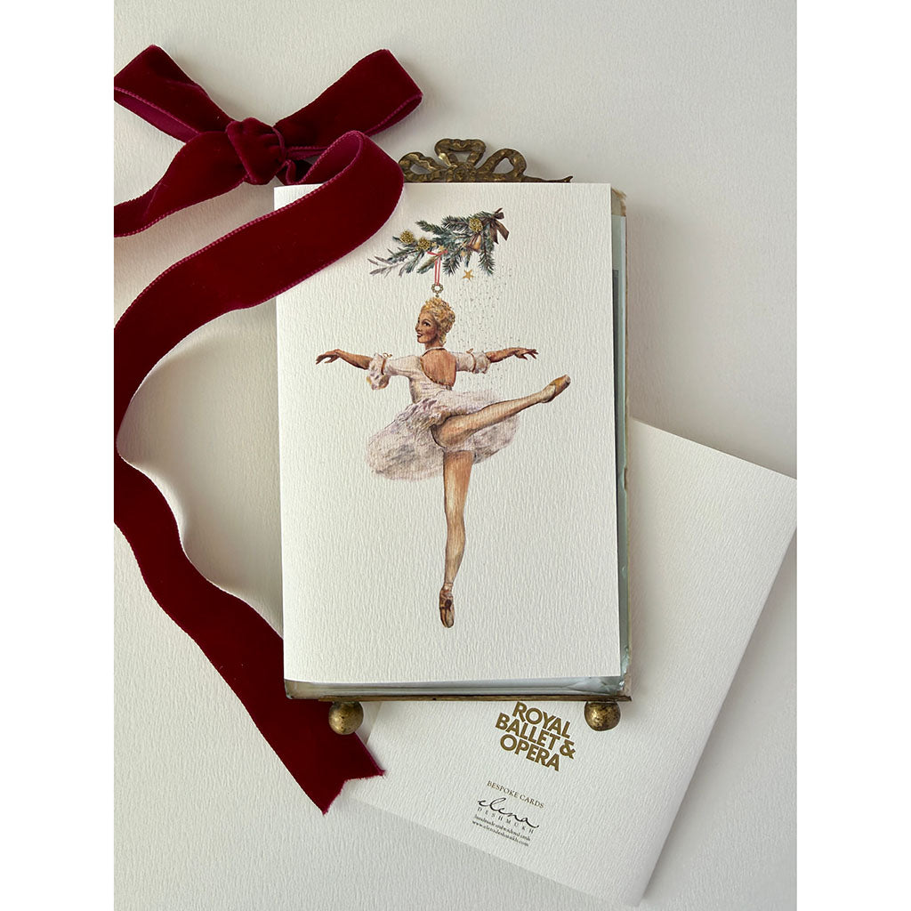 A handmade greeting card featuring a watercolour illustration of a ballerina in a white tutu suspended from a decorated Christmas tree branch, centred on a textured off-white background with a deep red velvet ribbon.