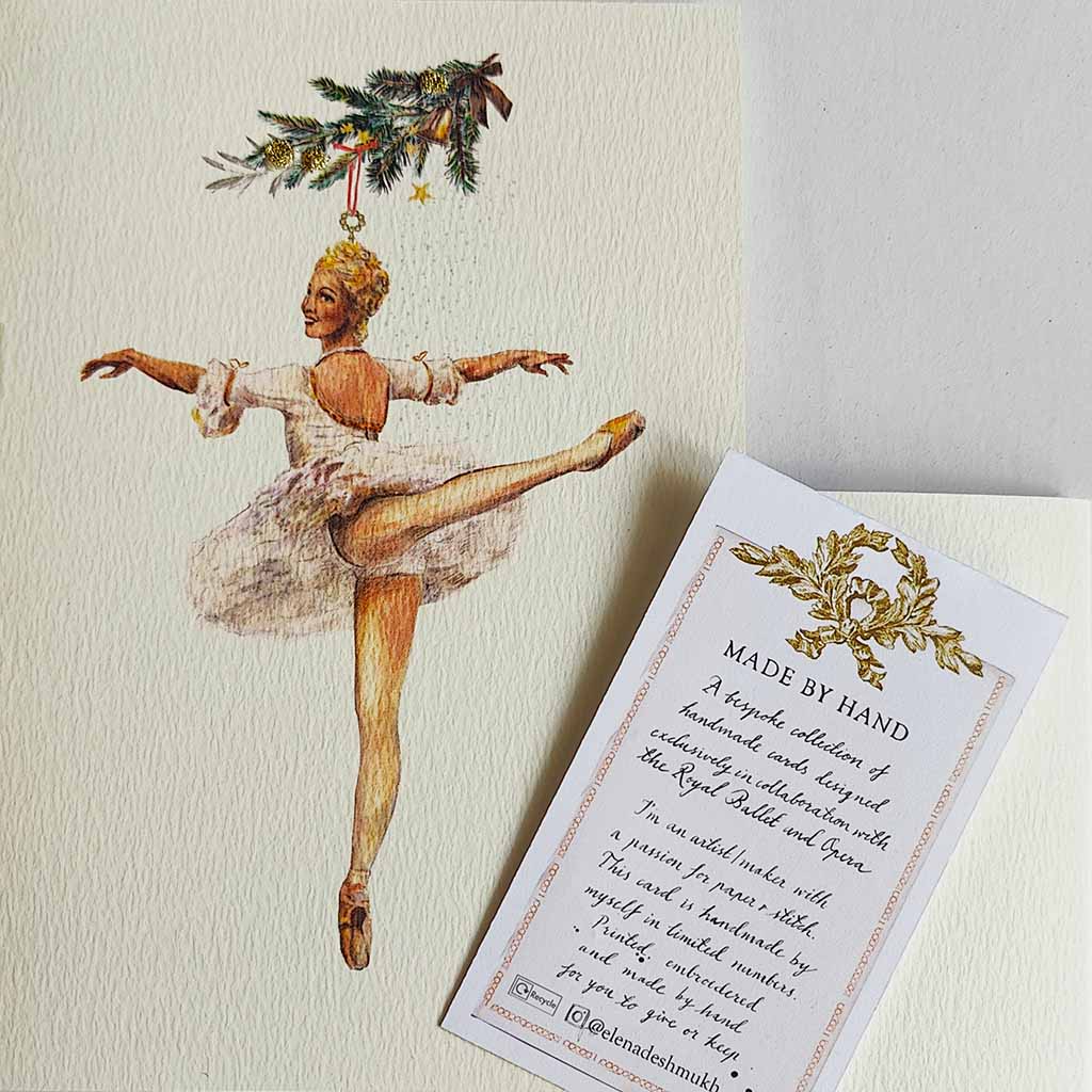A close up of a handmade greeting card featuring a watercolour illustration of a ballerina in a white tutu suspended from a decorated Christmas tree branch, centred on a textured off-white background.