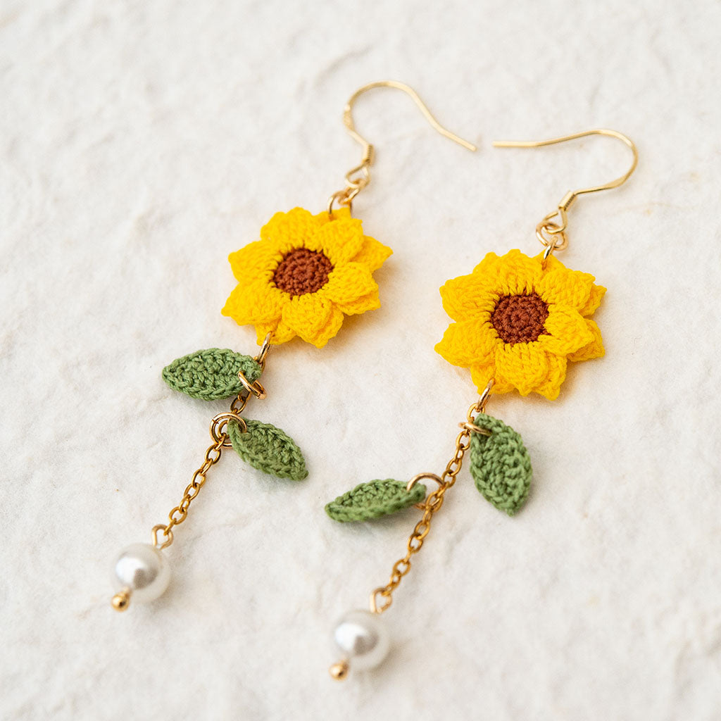 micro crochet sunflowers on gold drop earrings with leaves and pearls on a textured white surface