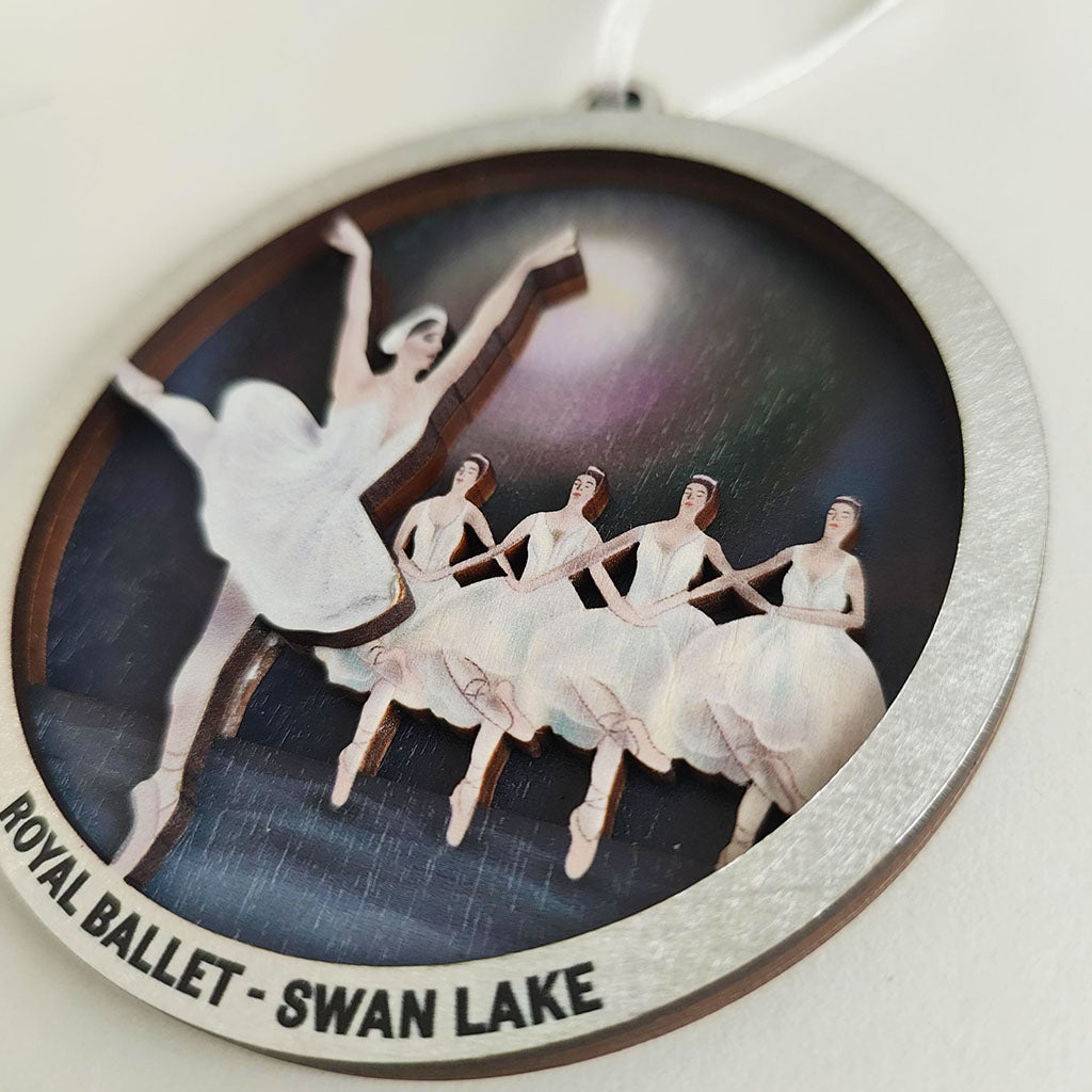A close up of a  round hanging decoration featuring a ballet illustration from Swan Lake, with ballerina figures in white tutus against a dark stage-like background and the text 'ROYAL BALLET - SWAN LAKE' around the edge.