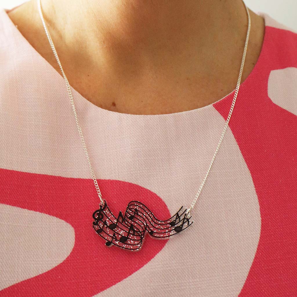 A close-up of a person wearing a silver necklace with a musical note and staff pendant, paired with a pink and white patterned top.