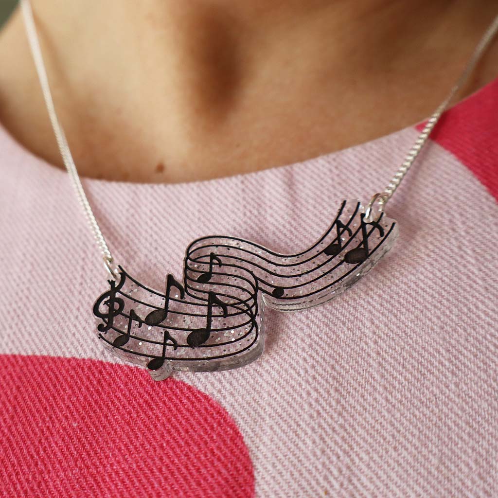 A close-up of a person wearing a silver necklace with a wavy musical staff and black music notes pendant, glittery background styled over a pink and red patterned top.