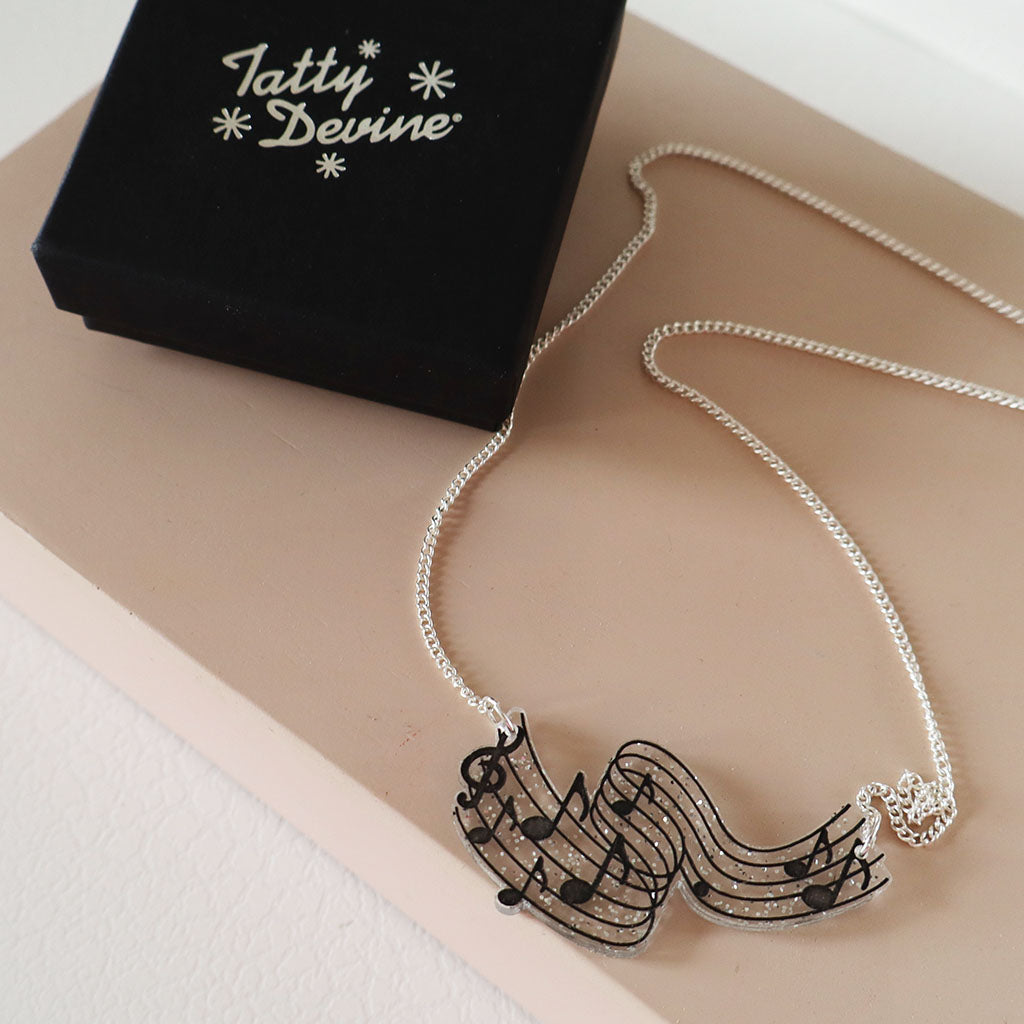 A silver chain necklace with a music note and staff pendant, displayed on a beige surface next to a black gift box printed with the name 'Tatty Devine'.