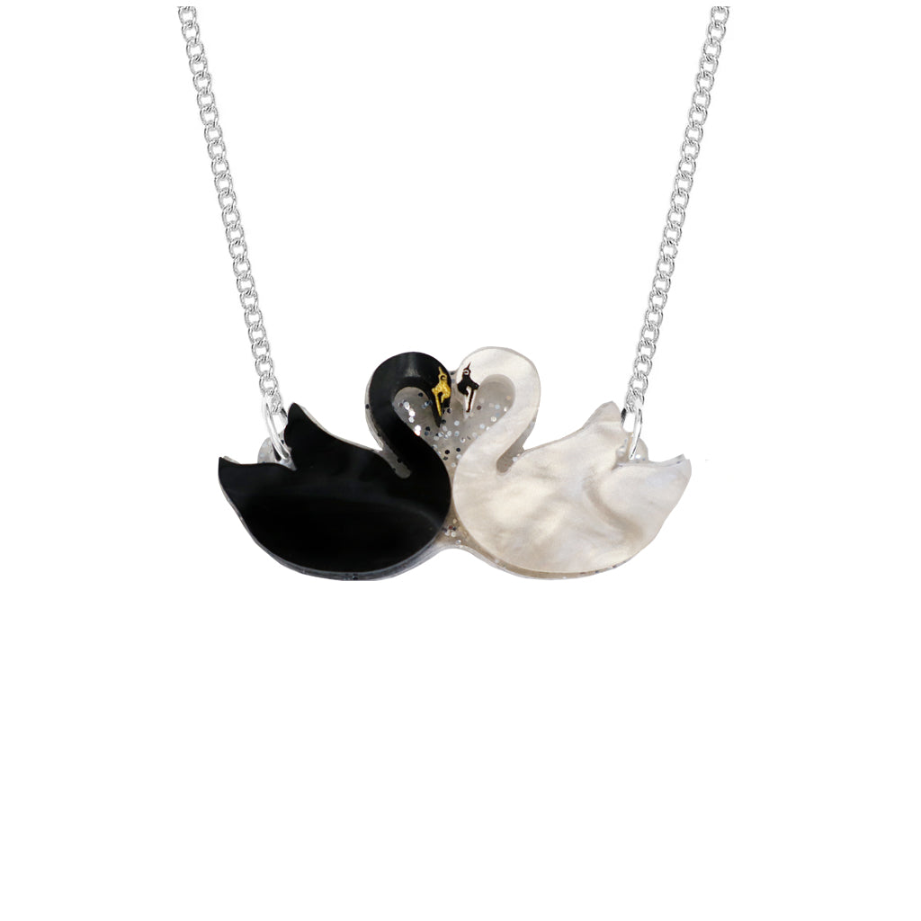 A silver chain necklace featuring a pendant shaped like two swans facing each other. One swan is glossy black, and the other is white with a textured, mother-of-pearl-like finish.
