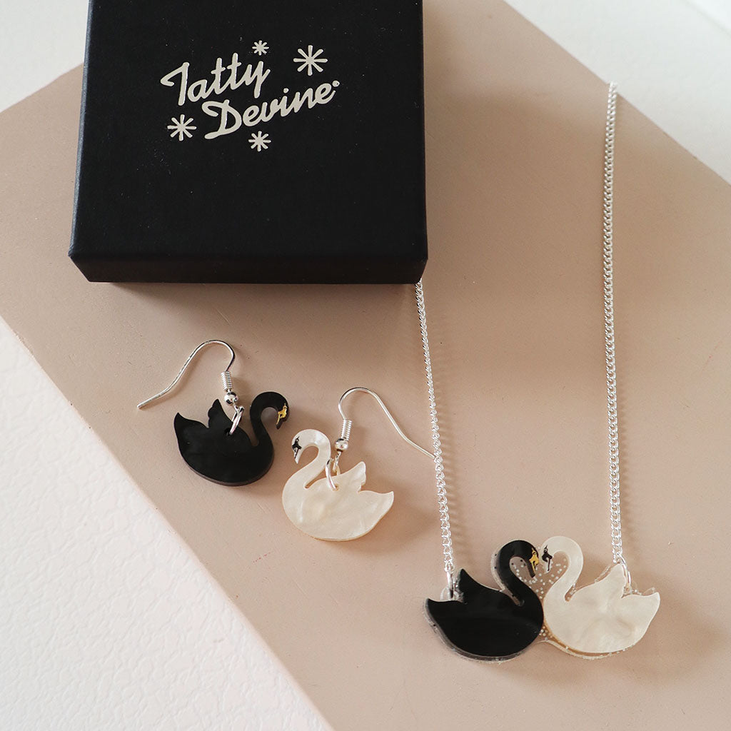 A pair of black and white swan drop earrings displayed next to a matching black and white swan pendant necklace on a silver chain, with a black gift box branded 'Tatty Devine' in the background.