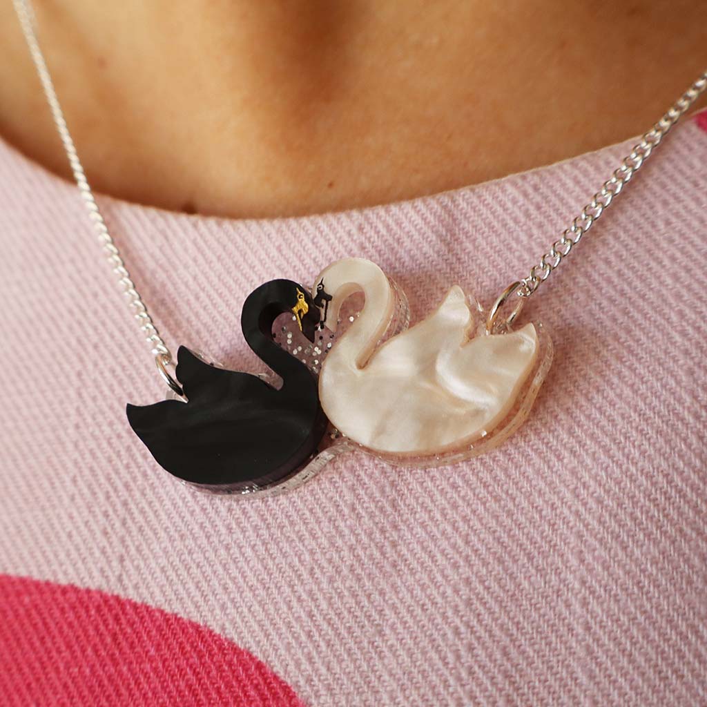 A silver chain necklace featuring a pendant shaped like two swans facing each other. One swan is glossy black, and the other is white with a textured, mother-of-pearl-like finish.