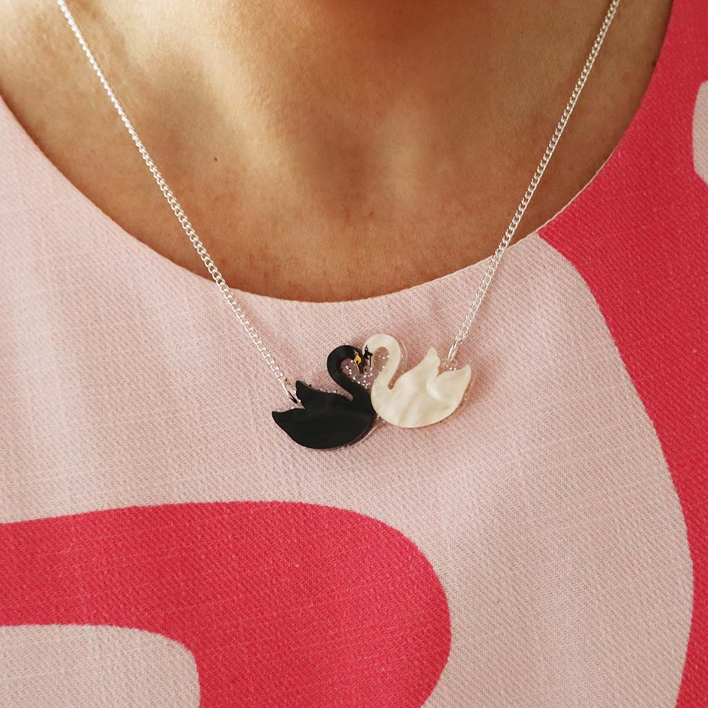 A close-up of a person wearing a delicate silver chain necklace with two swan pendants, one black and one white, facing each other. The person is wearing a pink and white patterned top.