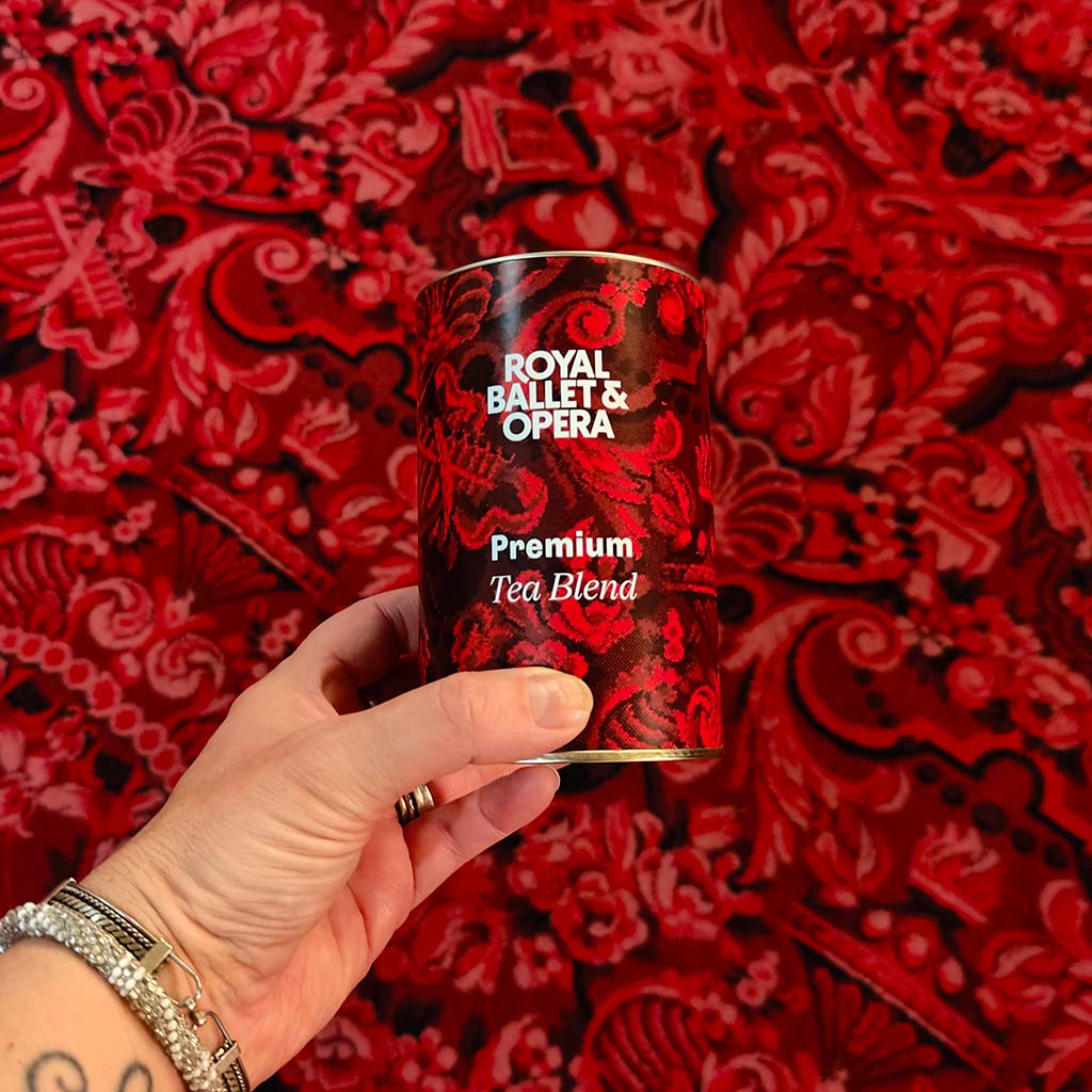 Hand holding a &#39;Royal Ballet &amp; Opera&#39; tea blend package against a red patterned background