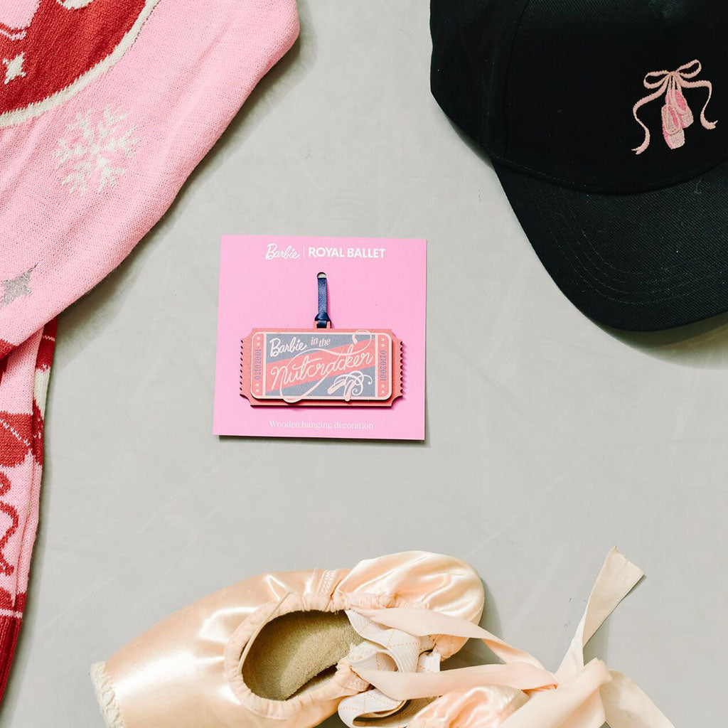 A flat-lay product photo of a 'Barbie Royal Ballet' Nutcracker Ticket Decoration on its pink backing card, styled with a pink Barbie Christmas Jumper, a black baseball cap with a pink ballet shoe graphic, and a satin pointe shoe on a light gray surface.