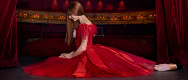 Marianela Nunez in a dramatic red gown sits on a dark stage floor in front of red curtains and a gold-trimmed backdrop.