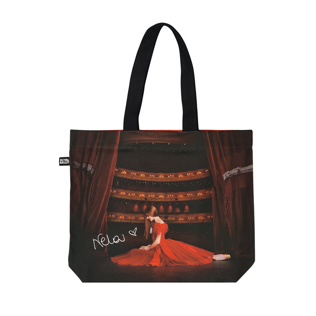 A tote bag featuring a printed graphic of ballerina Marianela Nunez in a red gown on a stage with red curtains, black handles, and a small black side tag with white text.
