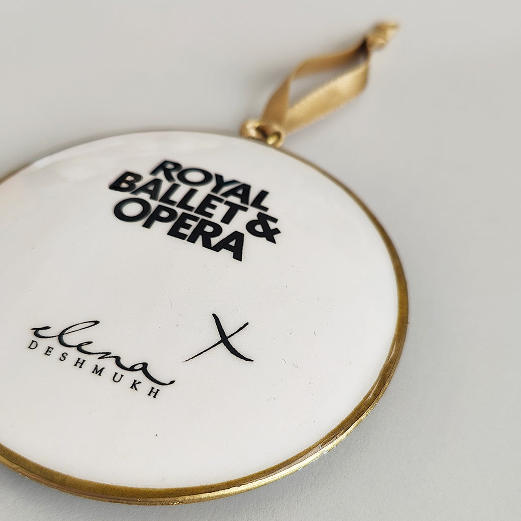 Close up of the design on the back of a circular tin decoration with black text Royal Ballet &amp; Opera Elena Deshmukh X on a white surface with a gold rim on a grey background