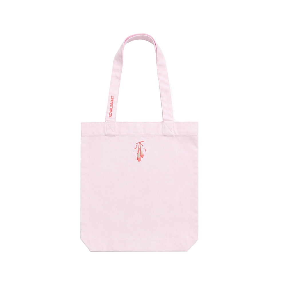 A light pink tote bag with matching pink shoulder straps, featuring a small embroidered illustration of ballet shoes in pink and coral near the centre, and the brand name 'ROYAL BALLET' printed vertically on the left strap.