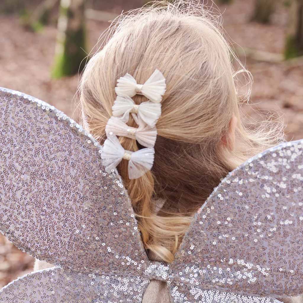 A close-up back view of a child with blonde hair styled in a braid, wearing four pink fabric bow hair clips. The child is also wearing a sparkly silver fairy costume with wings, set against a blurred outdoor forest background.