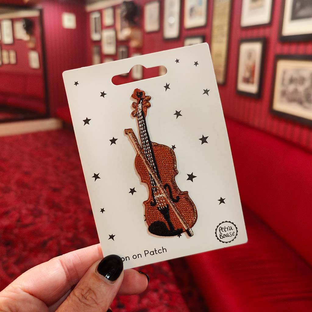 Embroidered violin patch on a card held in front of a red wall with framed pictures.