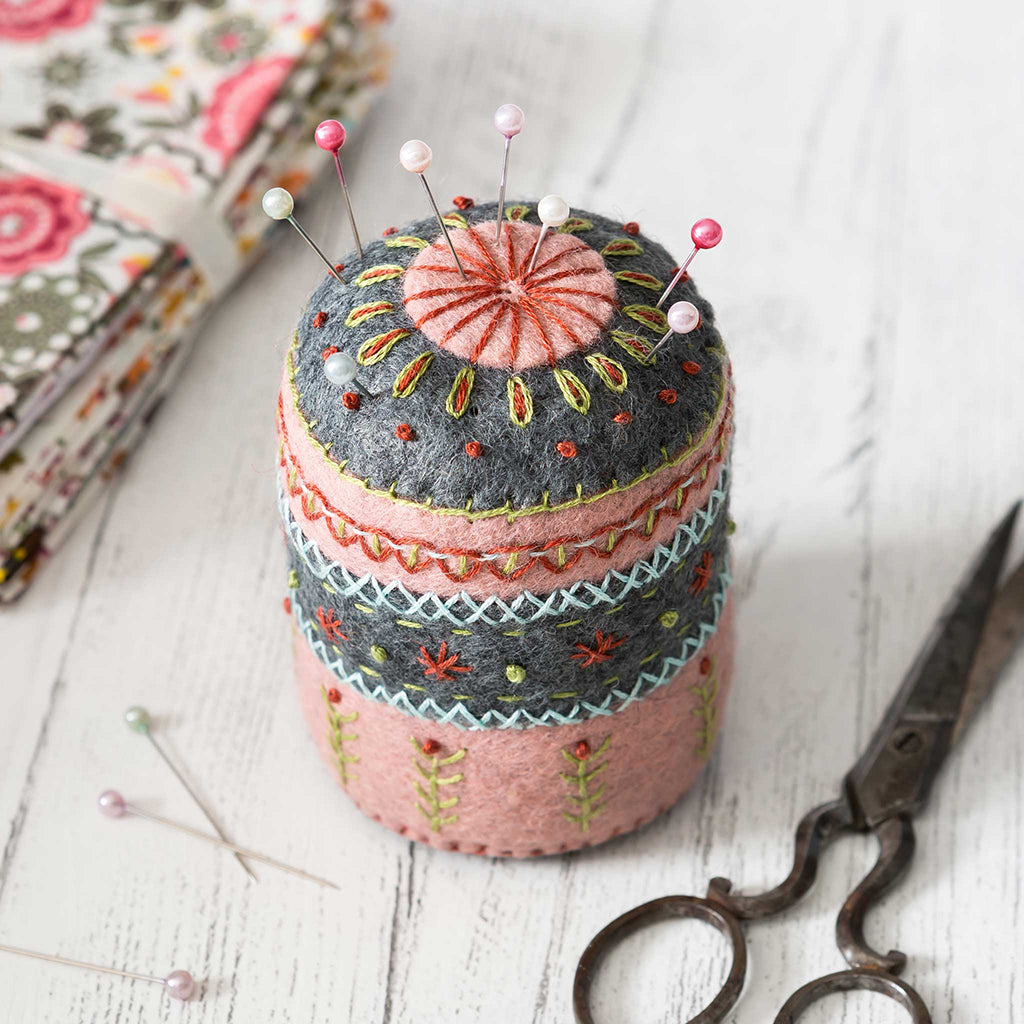 an embroidered felt pin cushion on a work bench with pins, scissor and fabric
